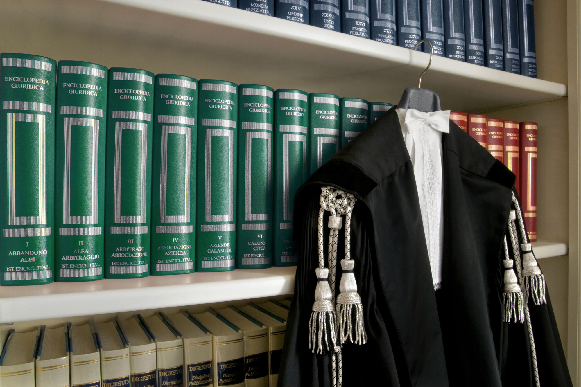 Toga nera da avvocato con cordoni argentati appesa davanti a volumi verdi dell'Enciclopedia Giuridica e del Digesto