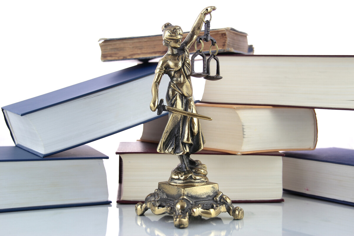 Statuetta in ottone della Giustizia bendata con bilancia e spada davanti a una pila di grossi libri dalle copertine blu e bordeaux
